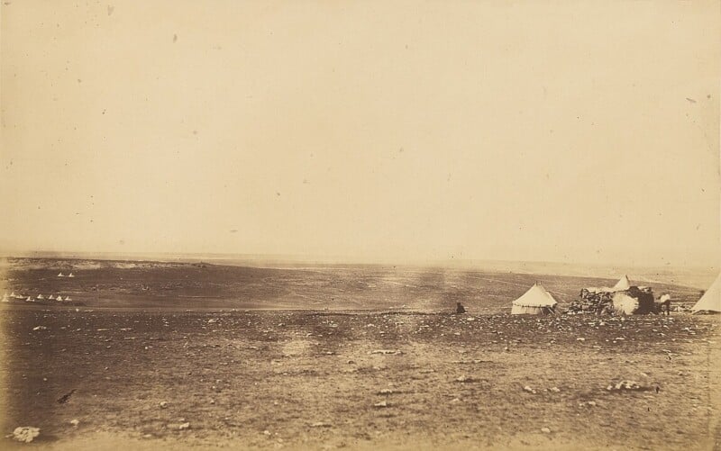 A sepia-toned image of an open landscape featuring a barren field with sparse vegetation. Two tents are visible on the right side, and a few more structures or tents are in the distant background under a wide, hazy sky.