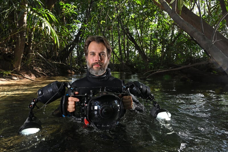 A man stands waist-deep in water, surrounded by lush green vegetation. He is wearing a black wetsuit and holding a large underwater camera with two attached lights. Sunlight filters through the trees, creating a natural, serene atmosphere.