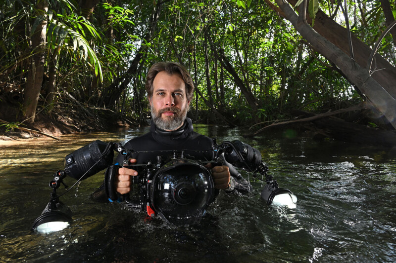 A man stands waist-deep in water, surrounded by lush green vegetation. He is wearing a black wetsuit and holding a large underwater camera with two attached lights. Sunlight filters through the trees, creating a natural, serene atmosphere.