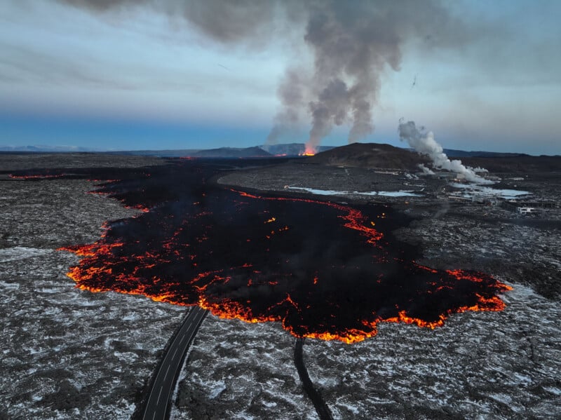 Aerial view of a lava field with glowing molten rock spreading across a landscape. Smoke rises from the active volcano in the background. A road runs through the foreground, partially surrounded by cooling lava. Industrial structures are visible nearby.