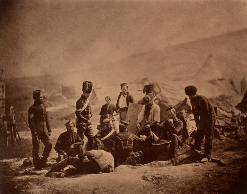 A sepia-toned historical photo shows a group of soldiers in uniform relaxing in a camp. They are gathered around a table with tents in the background. The scene conveys a moment of camaraderie during a military campaign.