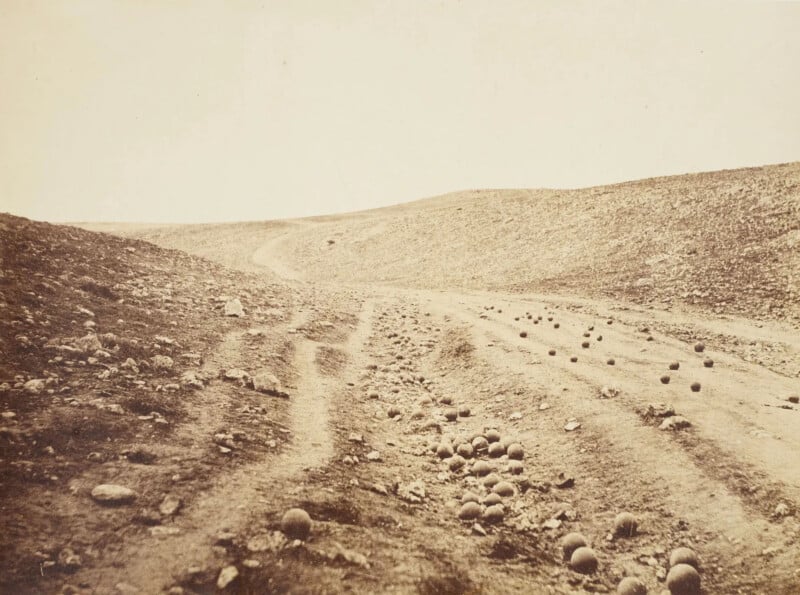 A sepia-toned photograph shows a barren battlefield landscape from the Crimean War. Cannonballs are scattered along a dirt road, which winds through a desolate and hilly terrain under a pale sky.