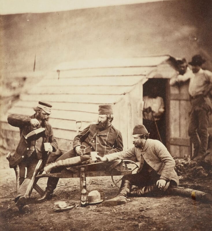 Three men in 19th-century military attire sit and drink around a wooden table outside a rustic cabin. Two pour drinks while one leans forward. Another man stands at the doorway, watching. A hat rests on the ground. The scene is sepia-toned.