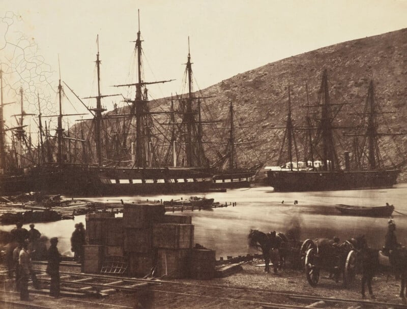 A sepia-toned historical photograph shows several large sailing ships docked in a harbor. The scene includes workers, horse-drawn carts, and stacked crates on the dock, set against a hilly backdrop.