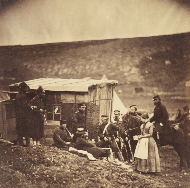 A group of people gathered outside a makeshift wooden structure and tent in a hilly landscape. Some are seated while others stand, dressed in 19th-century military uniforms and attire. A woman in a dress is among them, interacting with a soldier.