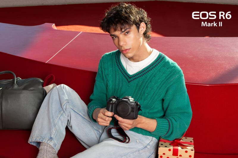 A person in a green sweater sits on a red couch holding a Canon EOS R6 Mark II camera. A gray bag and a wrapped gift with a red bow are next to them. "EOS R6 Mark II" is displayed in the top right corner.