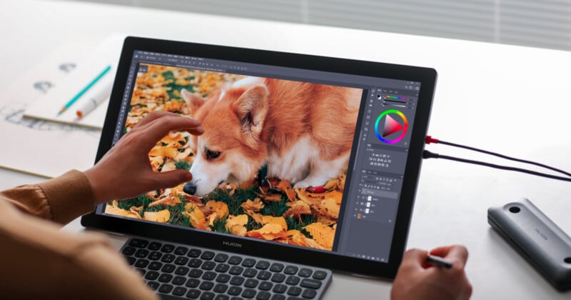 A person uses a stylus on a tablet displaying an image of a corgi among fallen autumn leaves. The hand gestures as if adjusting the picture. There's a graphics pen and paper on the desk.