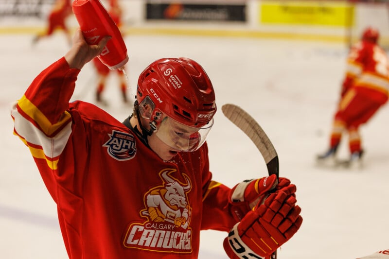 Hockey player in a red Calgary Canucks jersey holds a water bottle, gesturing with it while gripping his stick with the other hand. Other players are visible on the ice in the background.