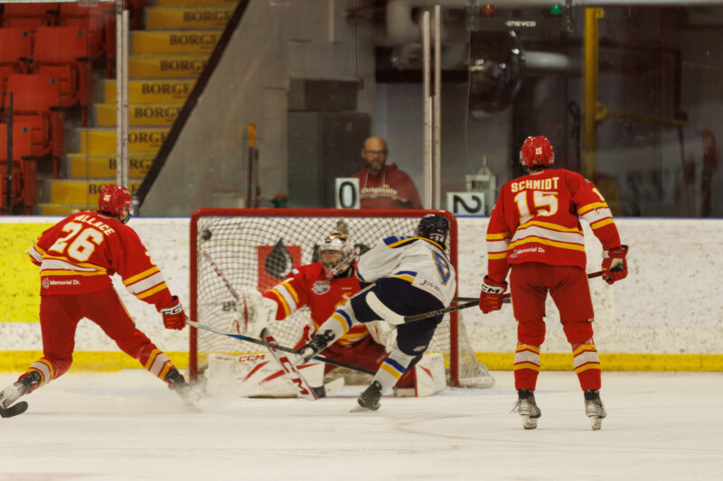 A hockey player in a blue and white uniform attempts a shot on goal, leaping in front of the red-clad goalie and two opposing players. The players and stands are visible in the background.