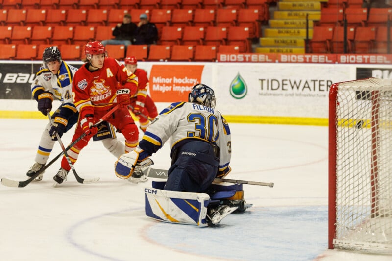 A hockey game in action: a player in a red jersey attempts to score as the goalkeeper in a blue and white jersey prepares to block the shot. Two other players, also in motion, are nearby on the ice rink. Red seating and advertisements are visible in the background.