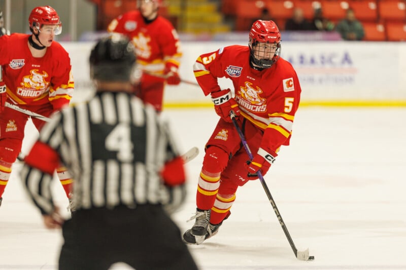 Hockey player in a red uniform with the number 5 skates near the referee during a game. Other players wearing matching jerseys are visible in the background on the ice rink.