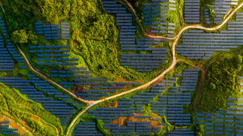 Aerial view of a vast solar farm on hilly terrain. Rows of blue solar panels are laid out in neat patterns, surrounded by lush green vegetation. A winding dirt path cuts through the landscape, connecting different sections of the solar farm.