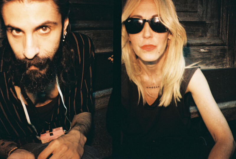 A bearded man in a striped shirt holds a camera while sitting next to a woman with long blonde hair, wearing sunglasses and a dark outfit. They are seated outdoors against a wooden backdrop.