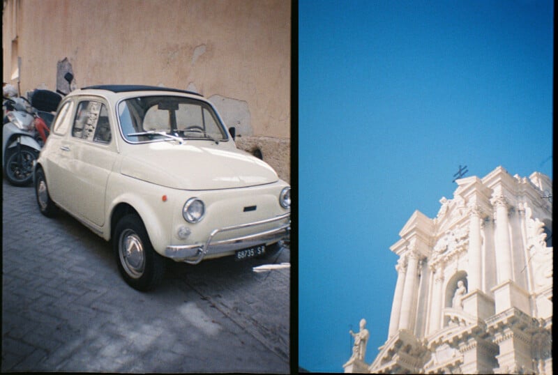 A vintage white car parked on a narrow street beside a wall and a scooter. To the right, a partial view of a classical building against a clear blue sky.