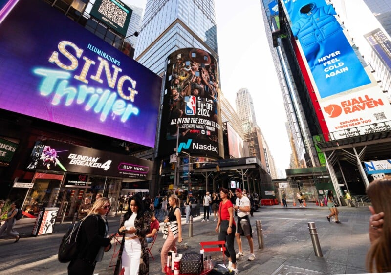 A bustling Times Square scene with bright billboards displaying ads, including "Sing Thriller" and an NBA ad. People walk and gather, enjoying the lively urban atmosphere under a clear sky.