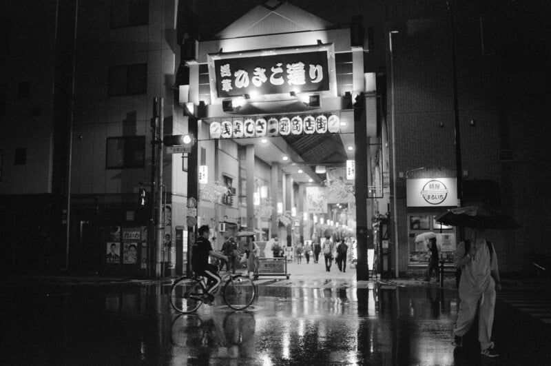 Photo en noir et blanc d’une scène de rue pluvieuse au Japon. Une entrée couverte de rue commerçante avec des panneaux lumineux en caractères japonais est visible. Une personne passe devant et une autre tient un parapluie. Les reflets scintillent sur le trottoir mouillé.