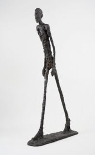 Une sculpture en bronze haute et élancée représentant un personnage marchant avec des membres allongés et une surface texturée, sur un fond blanc uni. La foulée du personnage est capturée au milieu du mouvement, transmettant mouvement et énergie.