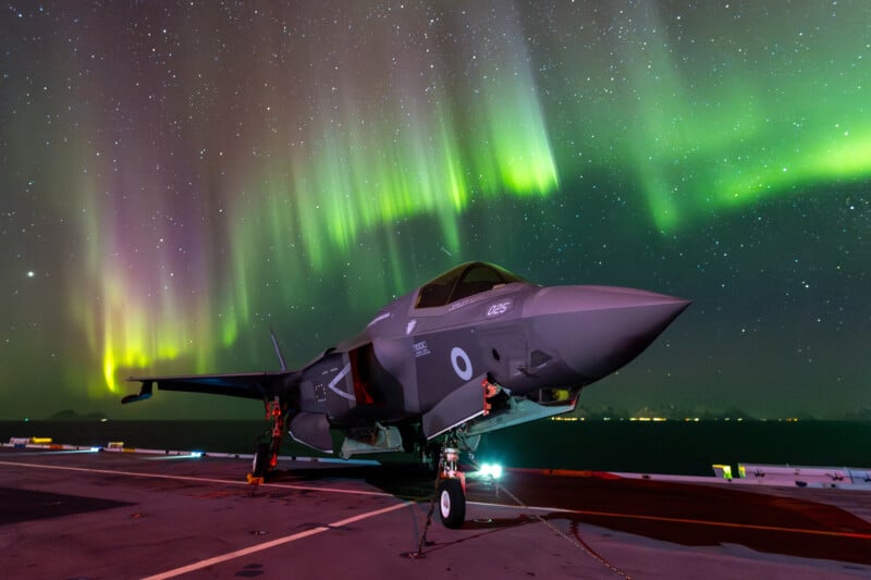 A fighter jet on the deck of an aircraft carrier under a vivid display of the Northern Lights. The sky is filled with green and purple auroras, creating a stunning backdrop against the night sky.