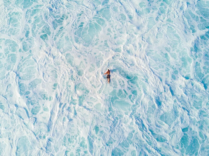 Aerial view of a person swimming in a vast ocean, surrounded by swirling white foam and turquoise waves. The water is bright and tumultuous, creating a striking contrast between the swimmer and the sea.