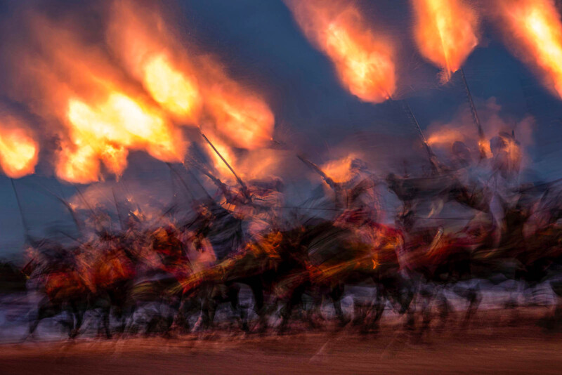 Blurred image of a group of riders on horses holding long poles with flames, creating a dramatic and dynamic scene against a dark blue sky. The motion blur adds a sense of speed and intensity to the fiery display.