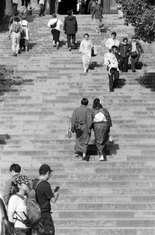 Des gens montent et descendent un grand escalier en pierre. Un couple en tenue traditionnelle se promène entre autres. Certaines personnes portent des sacs ou regardent leur téléphone. La scène est capturée en noir et blanc.