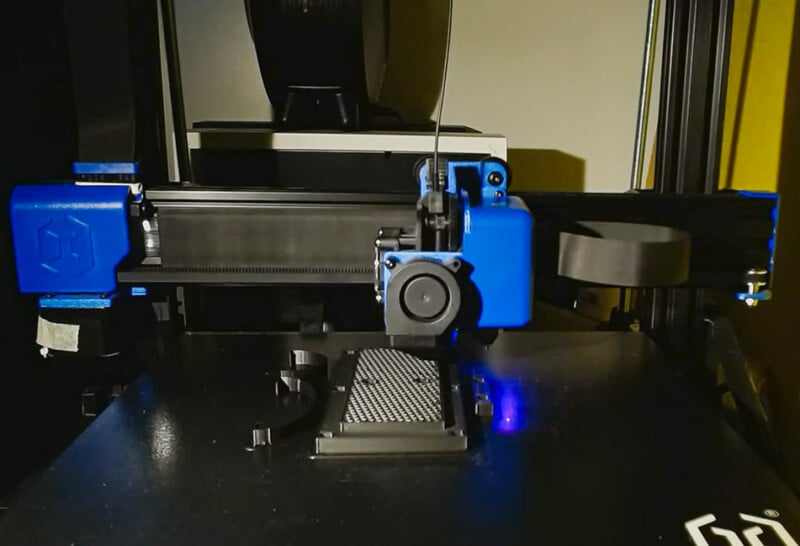 Une imprimante 3D avec des composants bleus imprimant un objet noir sur un plateau plat. Sa buse se déplace avec précision, déposant le filament couche par couche. La configuration est éclairée, révélant les détails complexes de la machine en action.