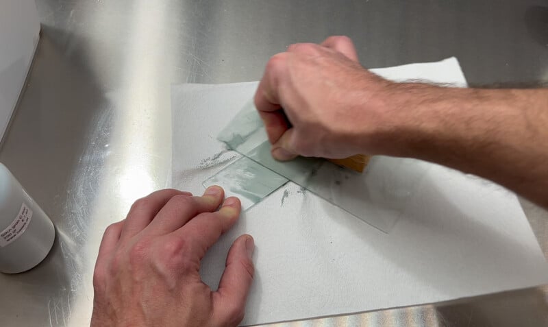 Une personne broie une substance sur une plaque de verre à l’aide d’une spatule. L'assiette repose sur une serviette en papier posée sur une surface métallique. Une petite bouteille est visible sur le côté gauche de l'image.