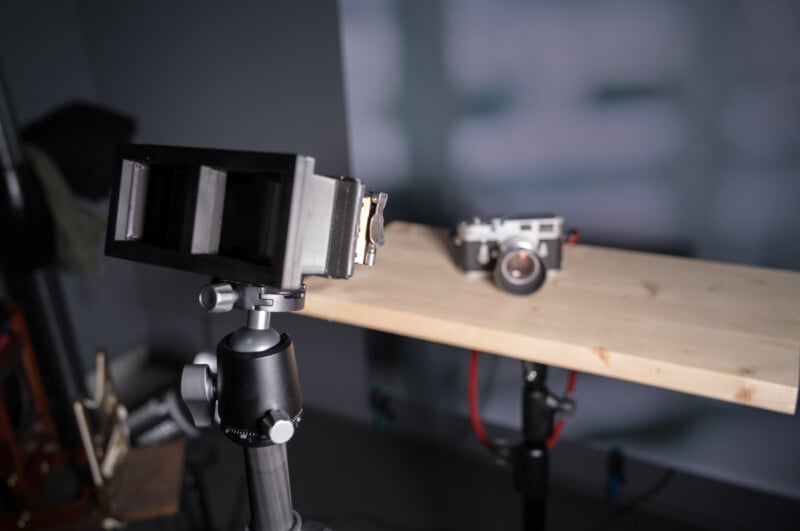 Un appareil photo vintage est monté sur une surface en bois devant un autre appareil photo posé sur un trépied. La configuration est à l’intérieur avec un éclairage doux et un arrière-plan flou.