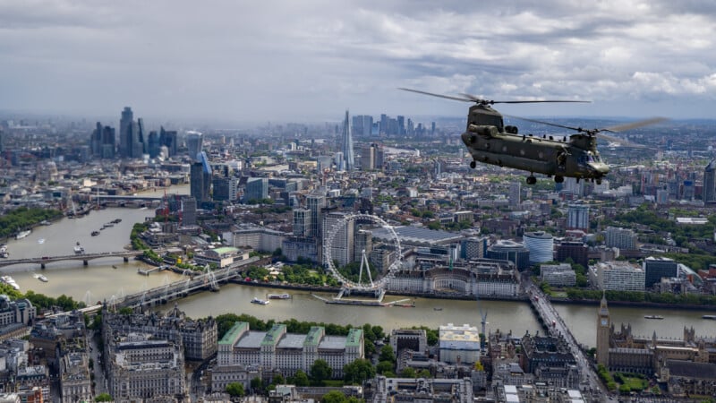 Aerial view of a cityscape with a military helicopter flying over a river. Notable landmarks include the London Eye and the Houses of Parliament. Skyscrapers are visible in the distance under a cloudy sky.