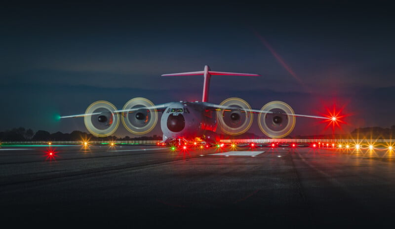 A large airplane with four propellers is stationary on a runway at night. The propellers are spinning, creating a circular light pattern. The runway is illuminated with red and white lights, and the sky is dark.