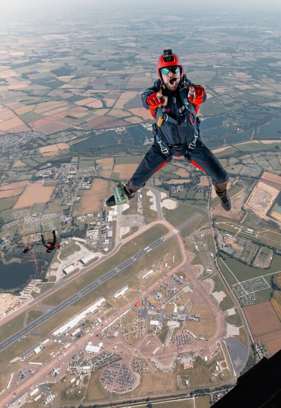 A skydiver in a red helmet and gear is mid-jump, making a joyful expression as they descend towards a landscape of fields and an airfield below. Another skydiver is visible in the background against the vast scenery.