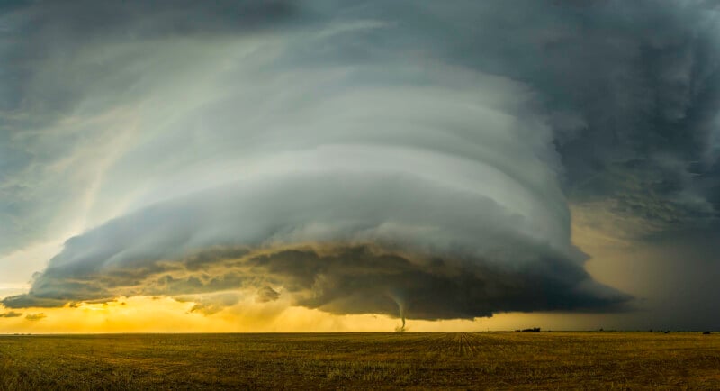 A dramatic scene of a supercell thunderstorm dominating the sky with swirling clouds and a visible tornado descending to the ground over a flat, open field. Dark, turbulent clouds contrast with a yellowish sky as sunlight peeks through.