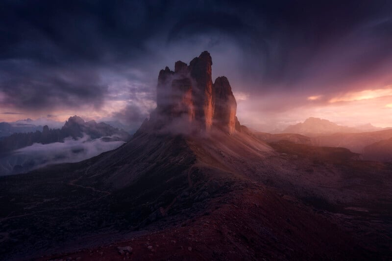 A dramatic mountain landscape at sunset, featuring towering rock formations shrouded in mist. The sky is heavy with swirling clouds, and the sun casts a warm glow on the peaks, creating a striking contrast with the surrounding shadows.