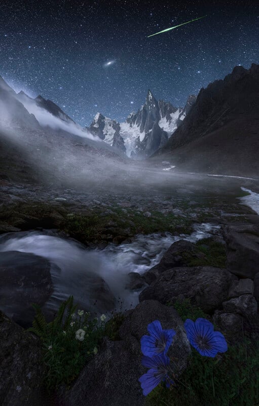 A serene mountain landscape at night with a starry sky and a meteor streaking above jagged peaks. A misty river flows through the foreground, surrounded by rocks and vibrant purple flowers.