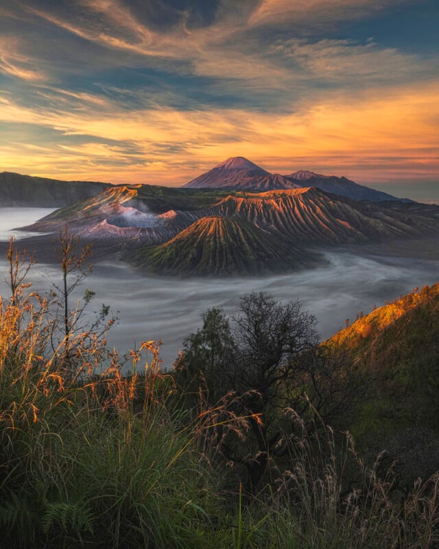 A scenic view of Mount Bromo at sunrise, showing the volcano surrounded by a sea of mist and rugged terrain. The sun casts warm, golden light on the landscape, with green vegetation in the foreground and a dramatic sky above.