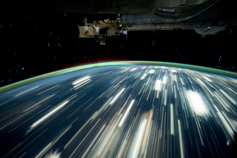 A stunning long-exposure photograph from space shows the Earth with vibrant, streaked lights across its surface. The curvature of the planet is visible against a dark, starry background, with part of a spacecraft in view.