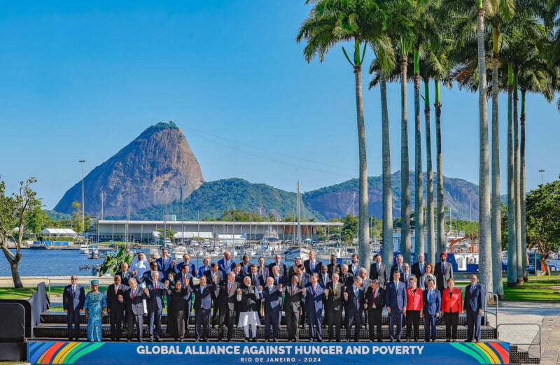 A group of people posing for a photo at the "Global Alliance Against Hunger and Poverty" event in Rio de Janeiro 2024. They stand on a stage with palm trees and Sugarloaf Mountain in the background.