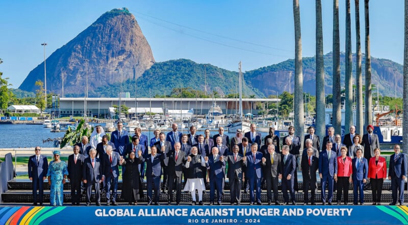 A large group of people in formal attire stand together at an outdoor event. A banner reads "Global Alliance Against Hunger and Poverty - Rio de Janeiro - 2024." A mountain and harbor with boats are visible in the background.