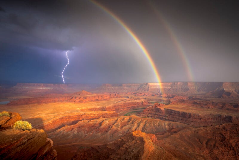 A dramatic landscape of a desert canyon under a stormy sky. A vivid double rainbow arches over the scene, with a bolt of lightning striking to the left. The canyon's reddish-brown rock formations are illuminated by soft, scattered sunlight.