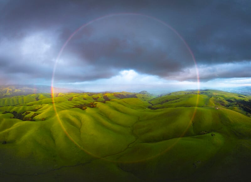 Aerial view of vibrant green rolling hills under a cloudy sky. A faint circular rainbow or halo is visible above the landscape, creating a serene and mystical atmosphere.