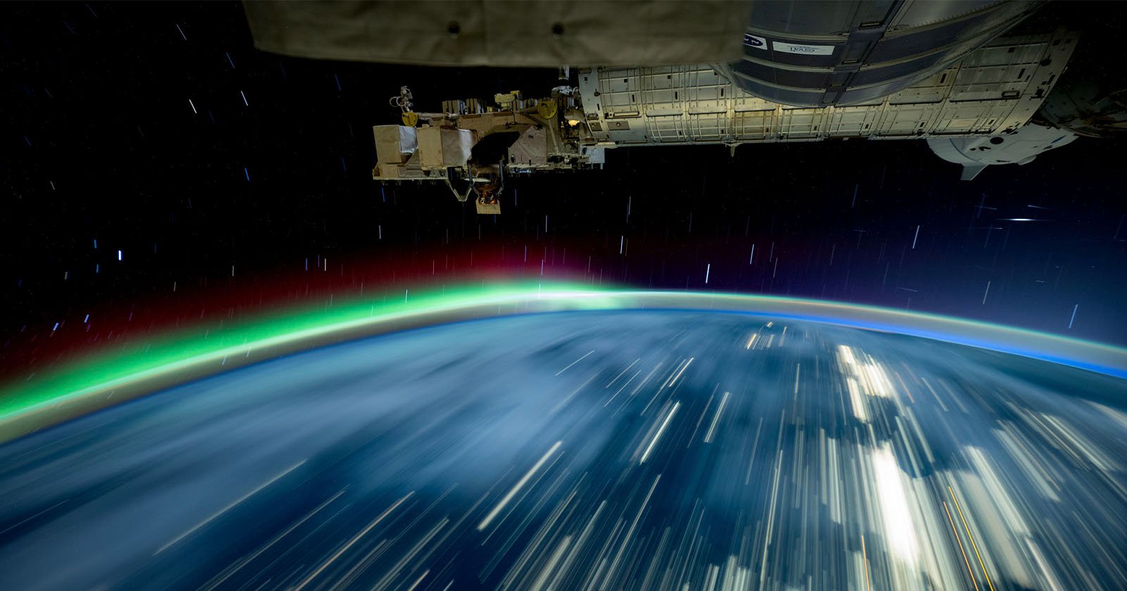 A long-exposure image taken from the International Space Station shows the Earth at night with vibrant streaks of aurora borealis in green and red. The blurred lights of cities contrast against the dark expanse of space.