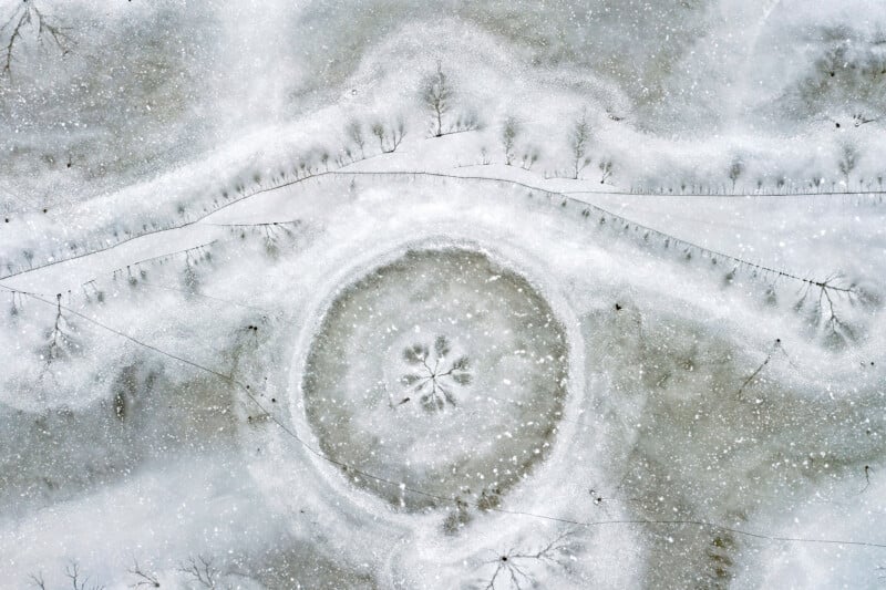 Aerial view of a snow-covered landscape featuring a large circular formation in the center, resembling a snow-covered pond or crater, surrounded by trees and faint paths. Snowflakes lightly cover the scene, creating a serene, wintry atmosphere.