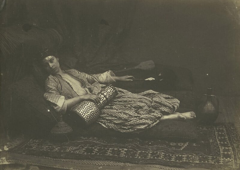 A person in traditional attire reclines on a patterned rug, holding a stringed instrument. They wear a headband and embroidered clothing, surrounded by ornate pillows and a decorative jug, creating a serene and nostalgic ambiance.