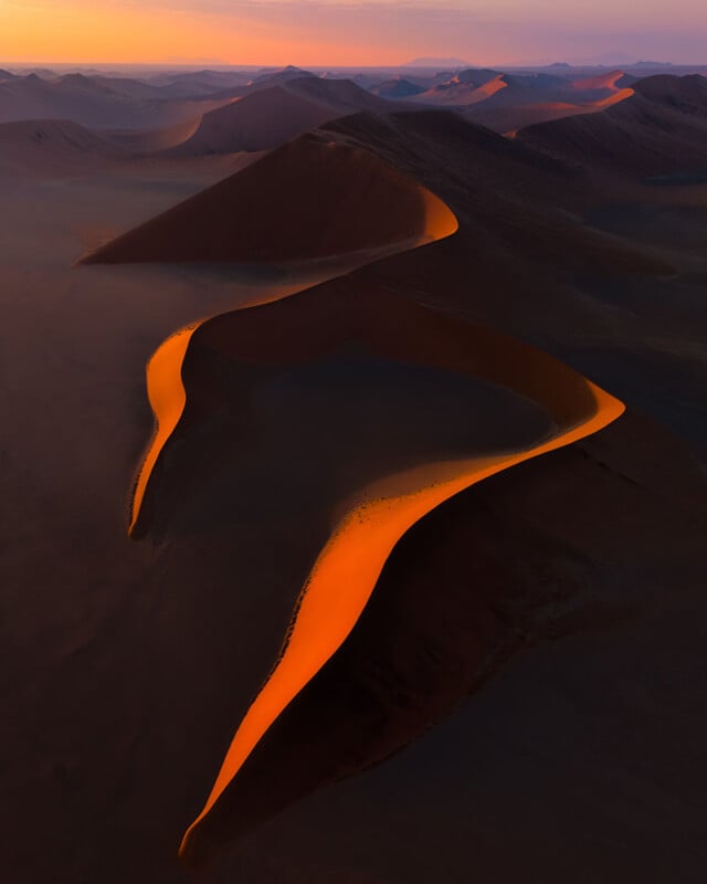 Aerial view of a desert landscape featuring a series of sand dunes illuminated by a warm, orange sunset. The light casts a striking contrast against the shadowy parts of the dunes, creating dramatic shapes across the scene.