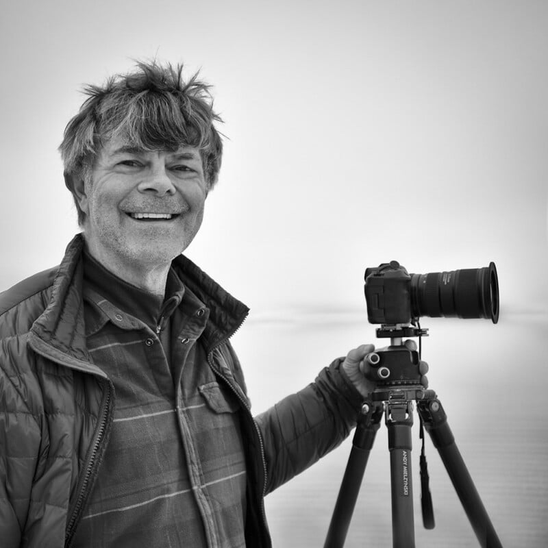 A smiling man with a windswept hairstyle stands beside a camera on a tripod. He is wearing a quilted jacket over a plaid shirt. The background is a soft, misty landscape. The image is in black and white.