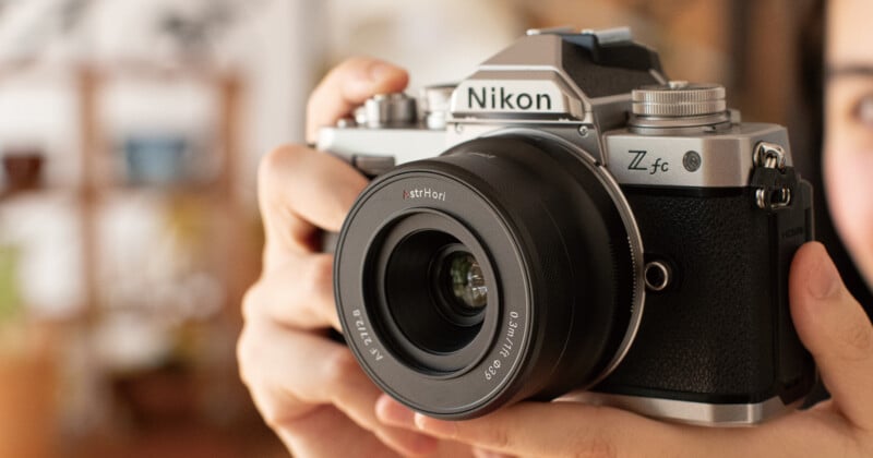 A person holding a Nikon Z fc camera with a 35mm lens. The camera has a vintage design with a silver and black finish. The background is softly blurred, highlighting the focus on the camera.