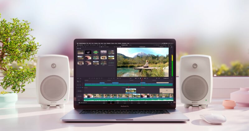 A laptop on a desk displays video editing software featuring a nature scene. Two white speakers are positioned on either side. The background includes a potted plant and blurred decor, suggesting a bright, modern workspace.