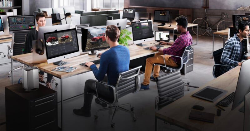 A modern office with four people working at desktop computers. The desks are arranged in two rows, with large monitors displaying graphics software. The office has an industrial design with an open layout and various tech equipment.