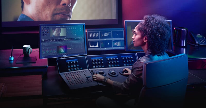 A person with curly hair sits at a high-tech editing console, working on multiple screens displaying video editing software, histograms, and color grading tools. The room is dimly lit with a modern and professional atmosphere.