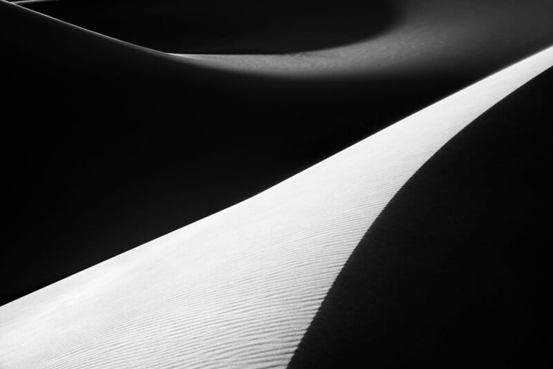 Black and white photograph of sand dunes with contrasting shadows and light. Smooth curves and textures are visible, highlighting the gentle slopes and patterns in the sand.